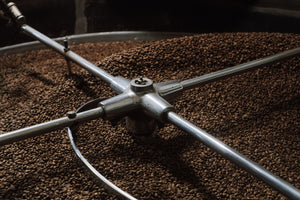 Close-up of coffee beans rotating inside a large metal roasting drum, with a four-spoke metal agitator arm crossing the frame.