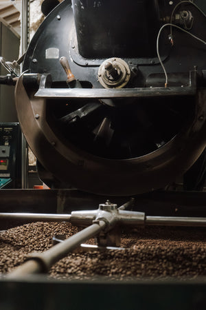 Close-up of a coffee roasting machine with a rotating drum and metal auger, with roasted coffee beans visible in the hopper below.